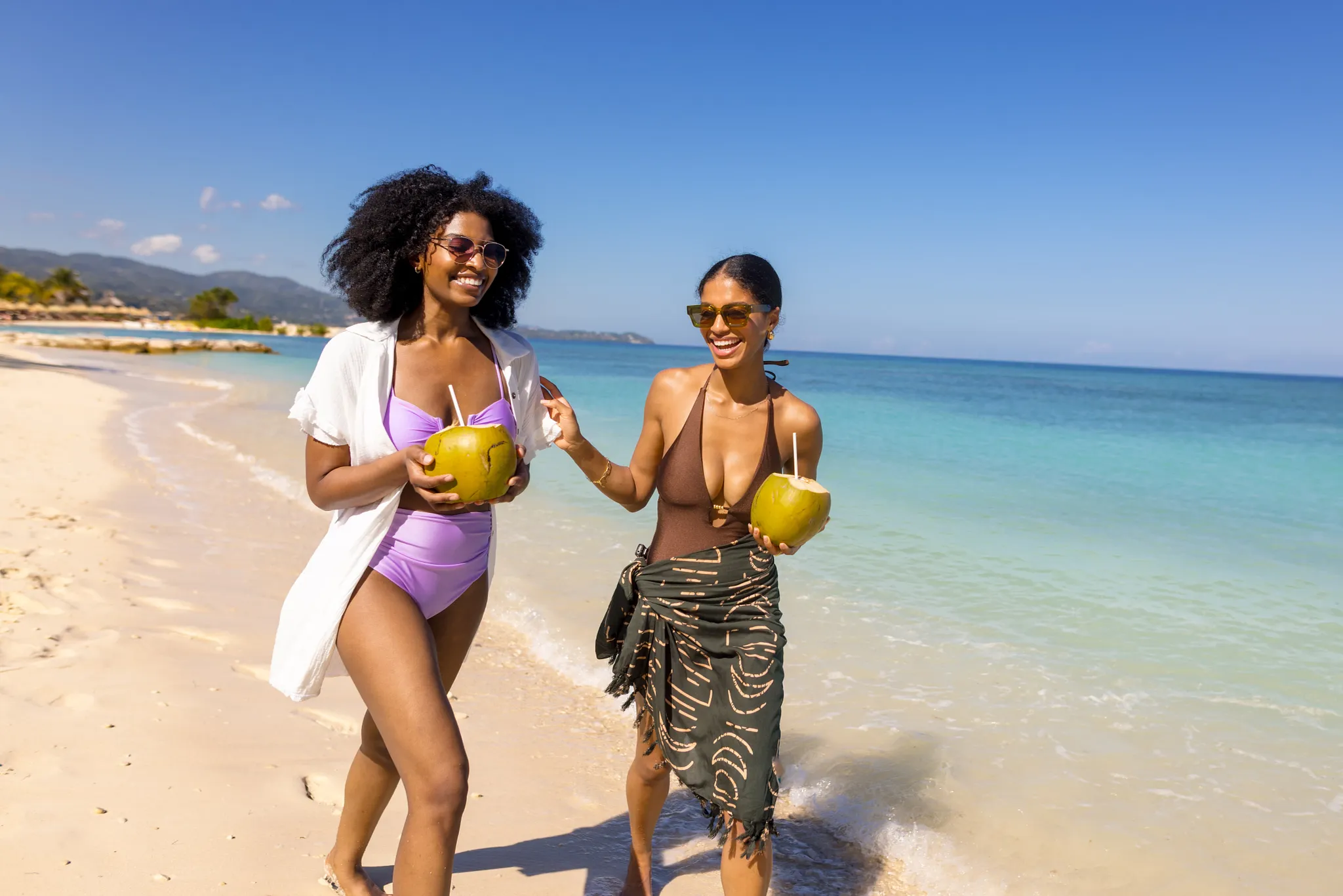 Two friends walking along the beach at Breathless Montego Bay holding fresh coconuts.